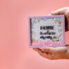 Hands holding a floral shadow box with 'Home is where mom is' text, perfect for Mother's Day gifting.